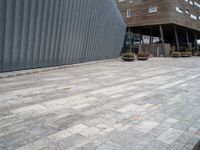 brick walkway next to an industrial building with wood trimming on the wall, and grey cement patio area, near planters and concrete sidewalk area with square seating area