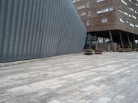 brick walkway next to an industrial building with wood trimming on the wall, and grey cement patio area, near planters and concrete sidewalk area with square seating area