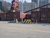 containers and equipment are on the ground near a street with buildings in the background and a crane is lifting them