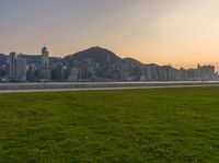 a city view with grass near a river and mountains as the sun sets over the horizon