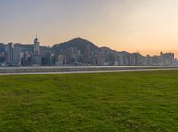 a city view with grass near a river and mountains as the sun sets over the horizon