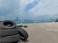 a pile of tires next to the ocean and city skyline with buildings in background, with the skyline with water and lots of buildings