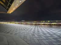 an empty walkway at night with bright lights on it and buildings in the background,