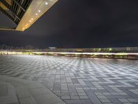 an empty walkway at night with bright lights on it and buildings in the background,