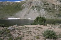 a horse in a field on the bank of a river near a mountain slope with a pond at the end and a mountain in the distance