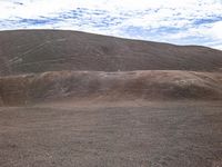 this person is riding a brown horse on a dirt field and is in the distance, with hills that are shaded by clouds