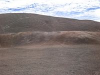 this person is riding a brown horse on a dirt field and is in the distance, with hills that are shaded by clouds