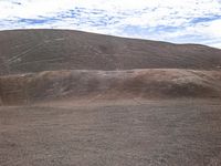 this person is riding a brown horse on a dirt field and is in the distance, with hills that are shaded by clouds