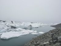 snow, ice, and rocks are in the water, with a person standing at a cliff overlooking the ice floes