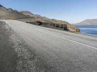 an empty road going into the distance between hills and a body of water with a person on it