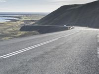 a car on a highway near the ocean and grassy area with water and hills in the background