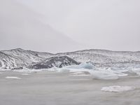 large number of ice pieces on the ground by water with snow covering it and mountains