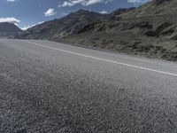 a motorcycle on the road surrounded by mountains and clouds in front of it, while someone is riding on one side of the lane