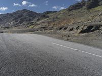 a motorcycle on the road surrounded by mountains and clouds in front of it, while someone is riding on one side of the lane