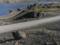 an automobile drives down the side of a winding road near the water and rocks, and a blue sky is in the background