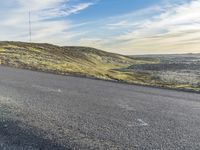 an empty road that is surrounded by grass and hills with a phone tower on top