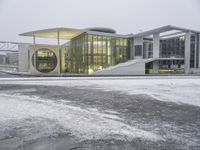 a bus drives in front of an impressive building in the snow, with lots of it's windows and concrete