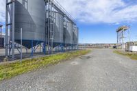 metal silos sit behind an industrial fenced yard with grass growing on each side