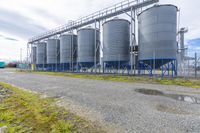 large steel silos standing on a gravel field near a large industrial building under a partly cloudy sky