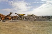an image of some very large machinery in the dirt near a hill of gravel, with construction cranes behind it