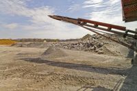 an image of some very large machinery in the dirt near a hill of gravel, with construction cranes behind it
