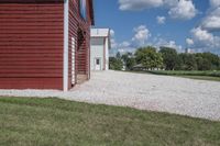 a barn on top of a gravel road next to grass and trees in front of a blue sky