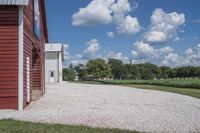 a barn on top of a gravel road next to grass and trees in front of a blue sky