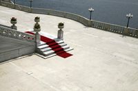 a red carpeting runner with pots of flowers on it near two white stairs overlooking a body of water