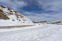 a dirt road covered in snow next to a rock formation and snowy hills with clouds