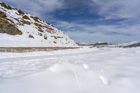 a dirt road covered in snow next to a rock formation and snowy hills with clouds