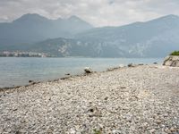 large rock and gravel beach next to body of water with mountains in background, during the day