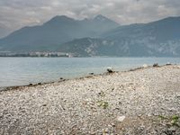 large rock and gravel beach next to body of water with mountains in background, during the day