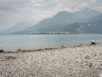 large rock and gravel beach next to body of water with mountains in background, during the day