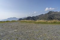 a mountain range with a rocky terrain covered with grass and plants near by, under a blue sky