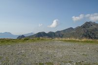 a mountain range with a rocky terrain covered with grass and plants near by, under a blue sky