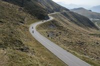 a long winding road cuts through the mountains, with a cyclist on it, riding down a curve