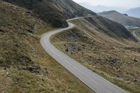 a long winding road cuts through the mountains, with a cyclist on it, riding down a curve