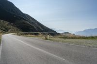 an empty asphalt road leading to a mountain range and some grassy hills with snow capped mountains in the distance