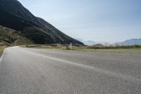 an empty asphalt road leading to a mountain range and some grassy hills with snow capped mountains in the distance