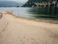 an empty beach on the shore of a mountain lake, with houses and seagulls in the background