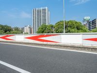 a large red and white arrow painted on a concrete wall by the road in front of a city with high buildings