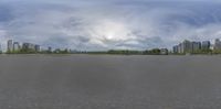 a wide empty road leading to some tall buildings and trees under clouds and sky is in view