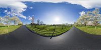 a small road is shown in a fish eye lens at the end of it there is an empty fence, bushes, and a sky with several clouds