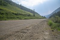 a dirt road and a hill side with trees on either side of it and a cloudy blue sky overhead