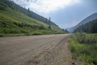 a dirt road and a hill side with trees on either side of it and a cloudy blue sky overhead