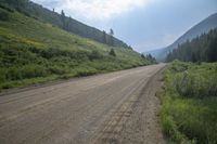 a dirt road and a hill side with trees on either side of it and a cloudy blue sky overhead