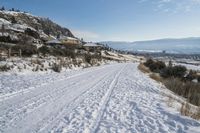 snow skiers on a snow covered road near a mountain with buildings and bushes on a hill