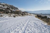 snow skiers on a snow covered road near a mountain with buildings and bushes on a hill