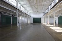 empty, open warehouse with metal poles and green doors and a concrete floor below a suspended ceiling