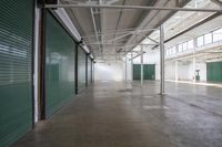 a long empty storage facility with green rolling doors on the wall and windows, and concrete floor